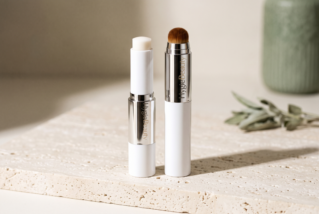 Les deux sticks du teint adaptatif 2-en-1 HygieBeauty, embout crème et pinceau intégré, sur une tablette de travertin.